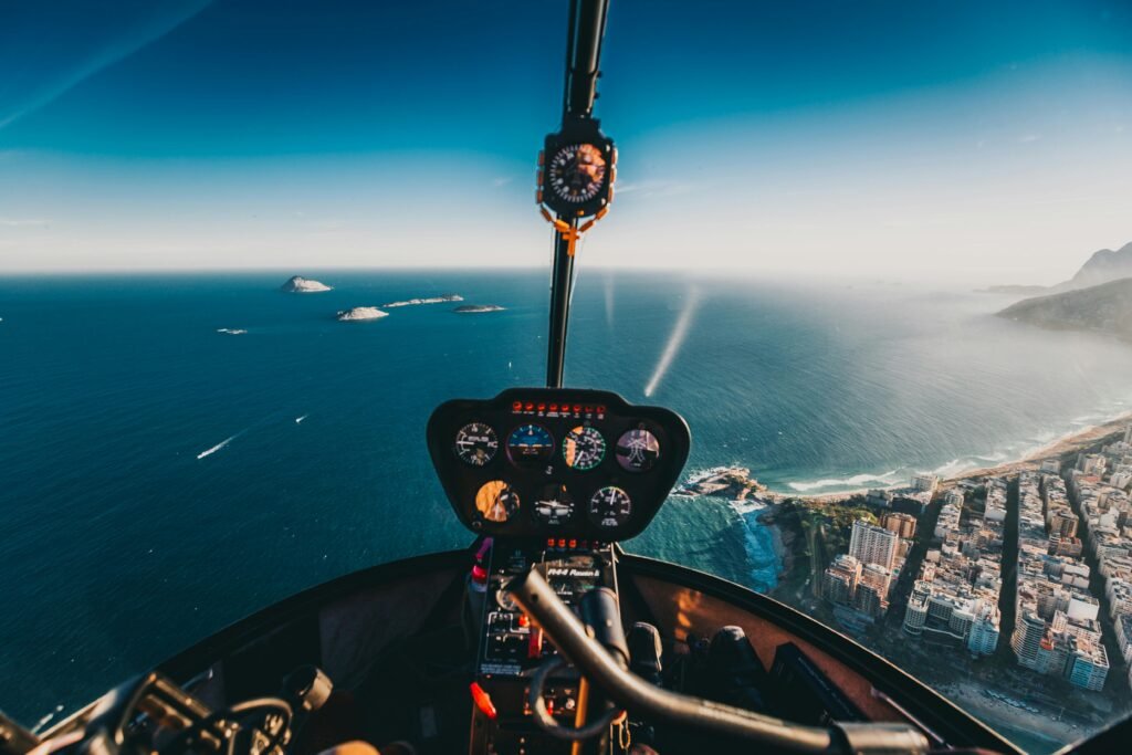 Cockpit view of a helicopter flying over the ocean with a coastal city and islands in the background.